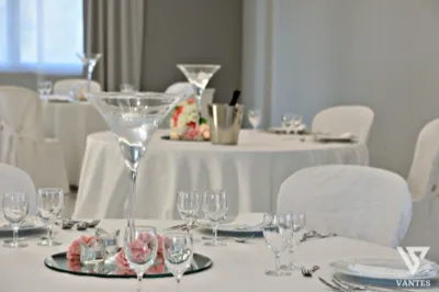 Dettaglio di una sala ricevimenti interna con tavoli rotondi rivestiti di bianco, bicchieri ordinati e sedie con coprisedia chiaro. Al centro del tavolo in primo piano c'è una grande coppa martini con candela galleggiante, accompagnata da piccoli portacandele e rose rosa su base a specchio.