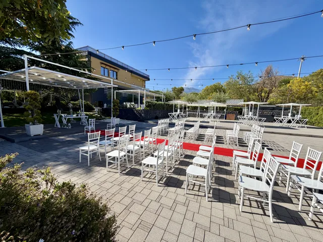 Area esterna allestita per rito simbolico di matrimonio con sedie bianche disposte su due lati, tappeto rosso centrale e tavolo cerimonia sotto una struttura coperta. Sullo sfondo si vedono tavoli alti, catenarie luminose e la villa, in una scenografia diurna ordinata e ariosa.