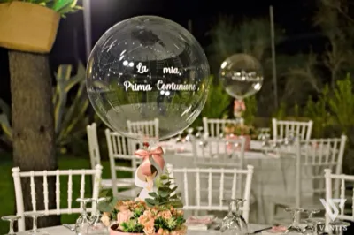 Dettaglio di un tavolo apparecchiato all'aperto di sera per una Prima Comunione, con sedie bianche, tovagliato chiaro e mise en place con nastri rosa. In primo piano spicca un centrotavola floreale con pallone trasparente personalizzato, mentre sullo sfondo si vedono altri tavoli coordinati nel giardino.