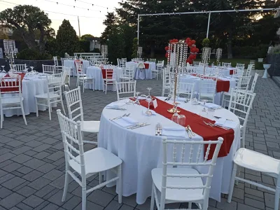 Allestimento esterno con tavoli rotondi apparecchiati in bianco e rosso su una grande pavimentazione, sedie bianche e alti centrotavola con cristalli pendenti. Sullo sfondo si vedono catenarie luminose, prato, alberi e un arco di palloncini che completa la scenografia della cerimonia.