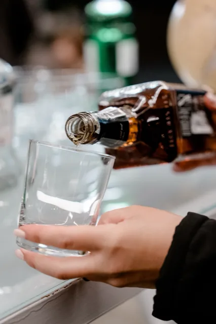 Primo piano di una mano che regge un bicchiere basso mentre da una bottiglia inclinata viene versato un amaro. L'immagine è molto ravvicinata e mette in evidenza il gesto del servizio drink durante l'evento.