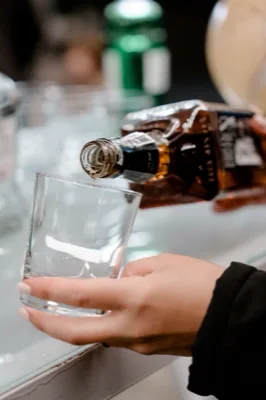 Primo piano di una mano che regge un bicchiere basso mentre da una bottiglia inclinata viene versato un amaro. L'immagine è molto ravvicinata e mette in evidenza il gesto del servizio drink durante l'evento.