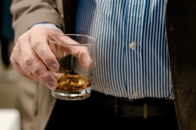 Primo piano di una mano che regge un bicchiere con un amaro ambrato, mentre sullo sfondo si intravede l'abbigliamento del servizio. La scena è ravvicinata e mette al centro il gesto della somministrazione del drink.