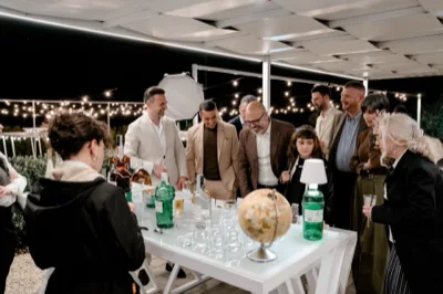 Gruppo di ospiti riunito attorno al banco amari all'aperto, con bottiglie, bicchieri e una luminosa area drink sotto la pergola serale. La scena mostra il momento conviviale del servizio, con persone che osservano e chiacchierano attorno al tavolo.