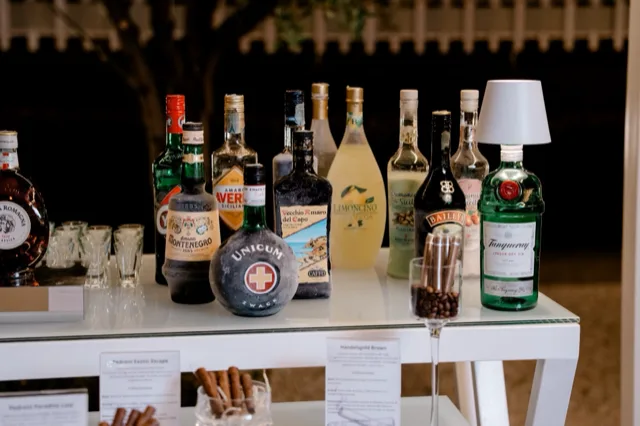 Primo piano del banco amari con numerose bottiglie esposte in fila, bicchierini trasparenti, cartellini descrittivi e piccoli contenitori con ingredienti o guarnizioni. L'inquadratura mette in evidenza la selezione di liquori e la presentazione curata del servizio beverage.