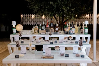 Banco amari allestito su più livelli all'aperto, con numerose bottiglie di liquori, bicchieri, cioccolatini e piccoli cartellini descrittivi ordinati sulla struttura bianca. La scena è illuminata di sera e mostra un servizio beverage ricco e molto curato.