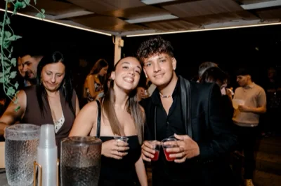 Area aperitivi all'ingresso con due giovani in primo piano che tengono bicchierini e sorridono verso la camera, mentre altri ospiti sostano dietro al banco beverage. Sul tavolo si vedono contenitori per drink, bicchieri e accessori da servizio sotto una copertura illuminata di sera.