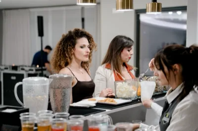 Buffet aperitivi con ospiti davanti al banco servizio, bicchieri, contenitori trasparenti con snack e una fila di drink colorati in primo piano. La scena è ambientata in una sala luminosa e mette in risalto il momento dell'aperitivo durante l'evento.