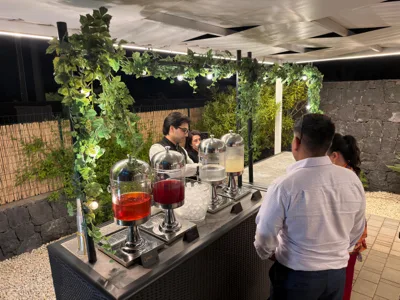 Postazione beverage all'aperto sotto una pergola decorata con fogliame, dove alcuni ospiti si avvicinano al banco per servirsi da grandi dispenser di bibite. La scena è illuminata di sera e mostra un momento di accoglienza o aperitivo nell'area esterna della location.