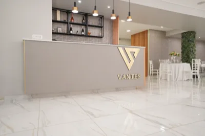 Bancone bar interno con logo Vantes illuminato sul fronte, sospensioni nere e oro sopra il piano e scaffalatura con bottiglie sul retro. Sullo sfondo si intravedono tavoli apparecchiati e dettagli della sala moderna con pavimento lucido e pareti dai toni neutri.