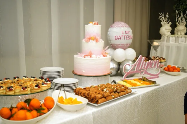 Tavolo dolci per battesimo con torta rosa a tre piani al centro, vassoi di cannoli, monoporzioni, frutta fresca e piatti pronti per il servizio. Sullo sfondo si vede un pallone decorativo con scritta dedicata al battesimo e il nome della festeggiata in evidenza sul tavolo.