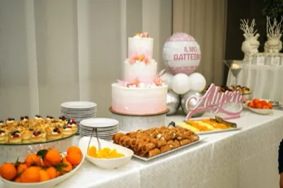 Tavolo dolci per battesimo con torta rosa a tre piani al centro, vassoi di cannoli, monoporzioni, frutta fresca e piatti pronti per il servizio. Sullo sfondo si vede un pallone decorativo con scritta dedicata al battesimo e il nome della festeggiata in evidenza sul tavolo.