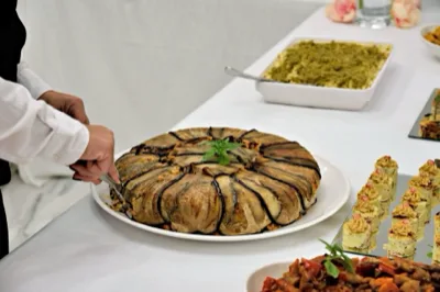 Grande timballo salato rivestito di melanzane servito su un tavolo buffet, mentre una persona in divisa lo porziona con le pinze. Sullo sfondo si vedono una teglia di pasta al forno e piccoli finger food disposti su vassoi da ricevimento.