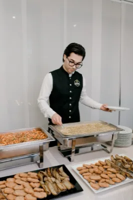 Cameriere al buffet caldo mentre serve primi piatti e fritti, con vaschette di pasta, piatti impilati e portate già pronte sul tavolo. In primo piano si vedono vassoi di fritti e contorni, in una postazione chiara e ordinata da servizio catering.