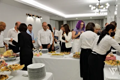 Buffet in sala con ospiti raccolti attorno ai tavoli delle portate, tra antipasti, formaggi, finger food e piatti pronti per il servizio. In primo piano e sul lato destro si vedono membri dello staff in divisa mentre assistono gli invitati durante il ricevimento.