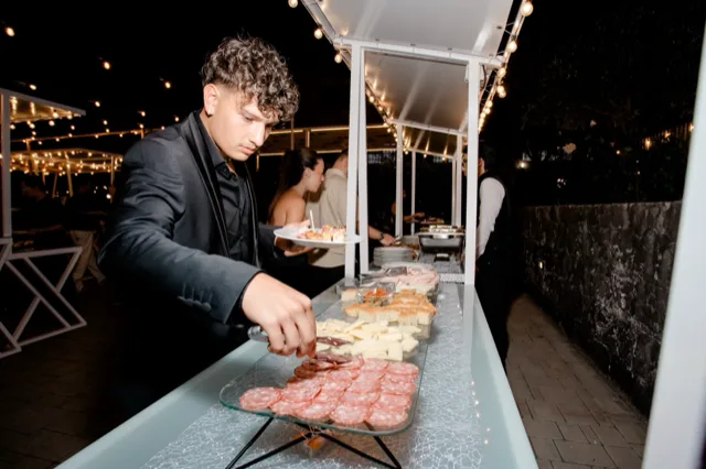 Buffet esterno serale con salumi, formaggi, panini e piccoli finger food disposti in fila sotto le luci calde della pergola. Un cameriere in abito scuro serve i piatti mentre gli ospiti si muovono lungo il banco all'aperto.
