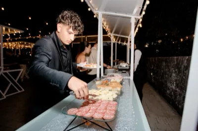 Buffet esterno serale con salumi, formaggi, panini e piccoli finger food disposti in fila sotto le luci calde della pergola. Un cameriere in abito scuro serve i piatti mentre gli ospiti si muovono lungo il banco all'aperto.
