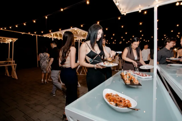 Buffet notturno all'aperto con un cameriere che serve salumi e formaggi da un lungo banco illuminato da catenarie calde. Sul tavolo ci sono vassoi di affettati, cubi di formaggio, pane, antipasti e piatti pronti, mentre gli ospiti si avvicinano lungo il percorso esterno.
