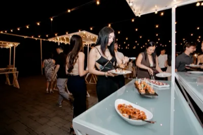 Buffet notturno all'aperto con un cameriere che serve salumi e formaggi da un lungo banco illuminato da catenarie calde. Sul tavolo ci sono vassoi di affettati, cubi di formaggio, pane, antipasti e piatti pronti, mentre gli ospiti si avvicinano lungo il percorso esterno.