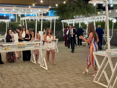 Ospiti eleganti che si muovono tra isole buffet all'aperto durante una serata, con tavoli bianchi illuminati e piatti già serviti in mano. Sullo sfondo si vedono pergole, alberi e catenarie luminose che definiscono l'area esterna del ricevimento.
