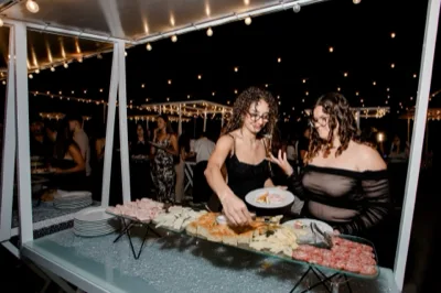 Buffet esterno notturno con due ospiti che si servono davanti al banco di salumi, formaggi, pane e antipasti, sotto le luci calde sospese nel giardino. Sullo sfondo si intravedono altri invitati e il servizio allestito sotto le pergole illuminate.