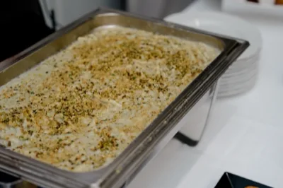 Vasca inox con pasta al forno o primo caldo servito a buffet, ricoperto da una spolverata di granella e spezie. L'immagine è un primo piano ravvicinato del piatto, con il tavolo bianco e la pila di piatti sullo sfondo.