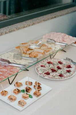 Tavolo buffet con salumi, formaggi, pane, tartine e piccoli assaggi monoporzione disposti su alzate di vetro e piatti bianchi. La scena è un primo piano molto ravvicinato dei vassoi e mostra una presentazione curata e ordinata da servizio catering.
