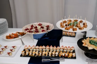 Tavolo buffet centrale con numerose portate fredde e calde disposte su alzate di vetro, piatti bianchi e vassoi neri. Si vedono finger food, salumi, formaggi, mini tramezzini e antipasti monoporzione in una presentazione ricca e ordinata da catering.