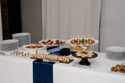 Tavolo buffet assortito con antipasti, finger food, tartine e salumi disposti su alzate di vetro, piatti neri e vassoi bianchi. Sul tavolo ci sono piatti impilati ai lati e un runner blu al centro che valorizza la presentazione delle portate.