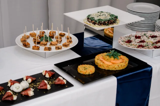 Buffet con portate monoporzione e finger food disposti su piatti neri e bianchi, inclusi tortini di riso, involtini, piccoli assaggi salati e un tortino decorato al centro. L'allestimento è ordinato, con tovaglia bianca e drappo blu sul bordo del tavolo.