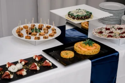 Buffet con portate monoporzione e finger food disposti su piatti neri e bianchi, inclusi tortini di riso, involtini, piccoli assaggi salati e un tortino decorato al centro. L'allestimento è ordinato, con tovaglia bianca e drappo blu sul bordo del tavolo.