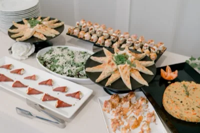 Tavolo buffet decorativo con triangoli di pizza o focaccia, involtini, piccoli tortini monoporzione, una ciotola di insalata e vassoi assortiti disposti su alzate nere e bianche. La presentazione è molto curata, con tovaglia chiara e drappo blu al centro.