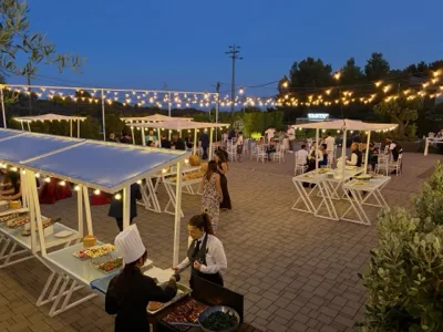 Vista serale dello spiazzale esterno con gazebo buffet illuminati, tavoli alti e ospiti distribuiti nell'area aperta sotto catenarie luminose. In primo piano si vedono personale di sala, postazioni catering e il grande piazzale organizzato per l'accoglienza e il servizio all'aperto.