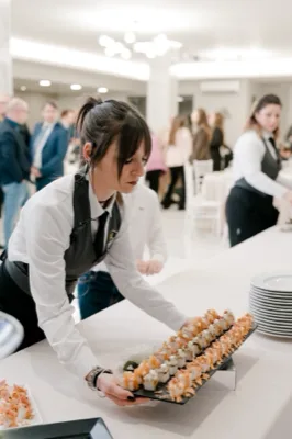 Cameriera al buffet che porge un lungo vassoio di finger food tipo sushi o canapè, in una sala luminosa con ospiti sullo sfondo. La scena è un momento di servizio ravvicinato, con piatti impilati e mise en place bianca ordinata.