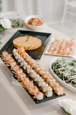 Tavolo buffet con file di finger food tipo sushi o canapè su vassoio nero, un tortino di riso al centro, insalata in ciotola bianca e altri piccoli assaggi disposti su alzate. La composizione è chiara, ordinata e molto ravvicinata, con stile catering elegante.