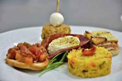 Piatto bianco con una selezione di antipasti misti serviti in piccole porzioni, tra cui una tartina con verdure, una crema salata decorata, un tortino giallo e una porzione gratinata. L'impiattamento è curato e ravvicinato, pensato per mostrare la varietà della proposta catering.