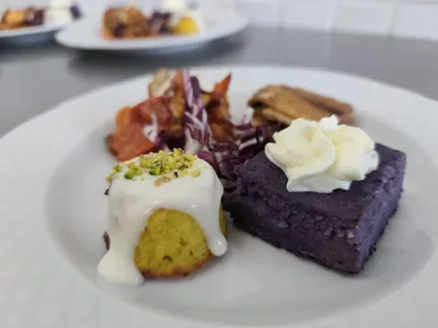 Primo piano di un piatto con piccoli assaggi gourmet, tra cui un tortino giallo con salsa bianca, una porzione viola con crema in superficie e altre preparazioni salate sullo sfondo. L'inquadratura ravvicinata mette in evidenza contrasti di colore, consistenze e cura dell'impiattamento.