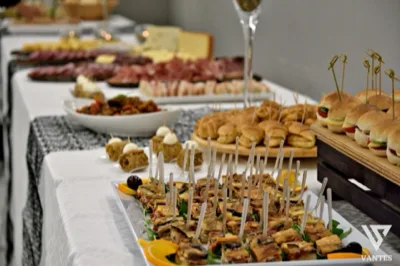 Tavolo buffet da apericena con finger food, mini panini, rustici e piccoli assaggi disposti in file ordinate su vassoi e alzate. Sullo sfondo si vedono salumi, formaggi e altre preparazioni fredde che completano una proposta ricca e curata.
