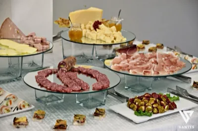 Buffet freddo con salumi, formaggi, miele e piccoli finger food disposti su alzate di vetro e vassoi bianchi in una presentazione ordinata. La scena mette in evidenza la varietà delle portate e la cura dell'allestimento per il servizio catering.