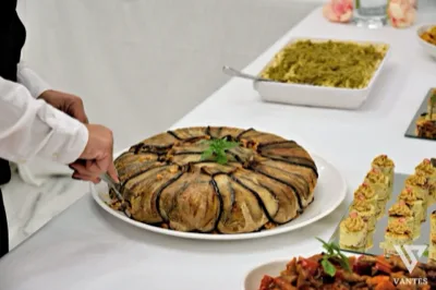 Grande timballo salato rivestito di melanzane servito su un tavolo buffet, mentre una persona in divisa lo porziona con le pinze. Sul lato destro si vedono finger food monoporzione e sullo sfondo una teglia di pasta e altre portate da ricevimento.