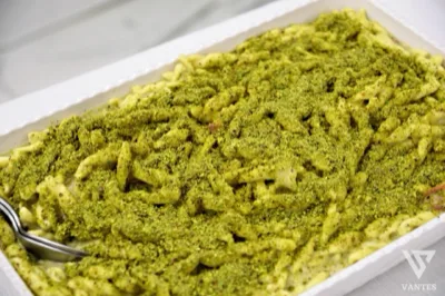Teglia di pasta corta condita in modo cremoso e ricoperta da una generosa granella verde, ripresa da vicino come proposta di primo piatto al buffet. L'immagine mette in risalto consistenza, condimento abbondante e presentazione da servizio catering.