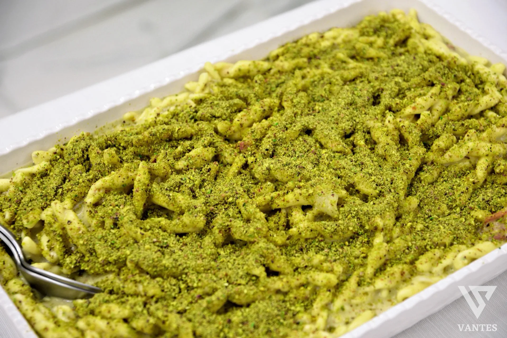 Teglia di pasta corta condita in modo cremoso e ricoperta da una generosa granella verde, ripresa da vicino come proposta di primo piatto al buffet. L'immagine mette in risalto consistenza, condimento abbondante e presentazione da servizio catering.