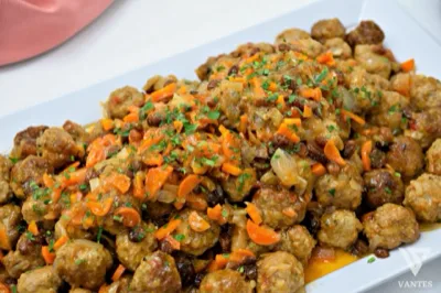 Grande vassoio di polpettine servite con un condimento ricco di cipolla, carote, uvetta e salsa lucida, ripreso in primo piano. La portata ha un aspetto generoso e casalingo, pensato per il servizio buffet di un secondo piatto caldo.