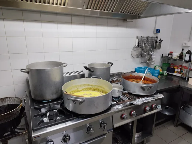 Cucina professionale con grandi pentole sui fuochi, salse in preparazione e utensili appesi alla parete in acciaio e ceramica bianca. La foto racconta un momento operativo del laboratorio catering durante la cottura delle portate.