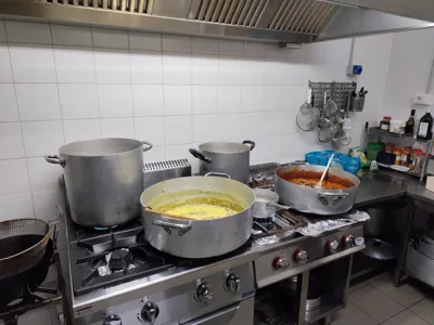 Cucina professionale con grandi pentole sui fuochi, salse in preparazione e utensili appesi alla parete in acciaio e ceramica bianca. La foto racconta un momento operativo del laboratorio catering durante la cottura delle portate.