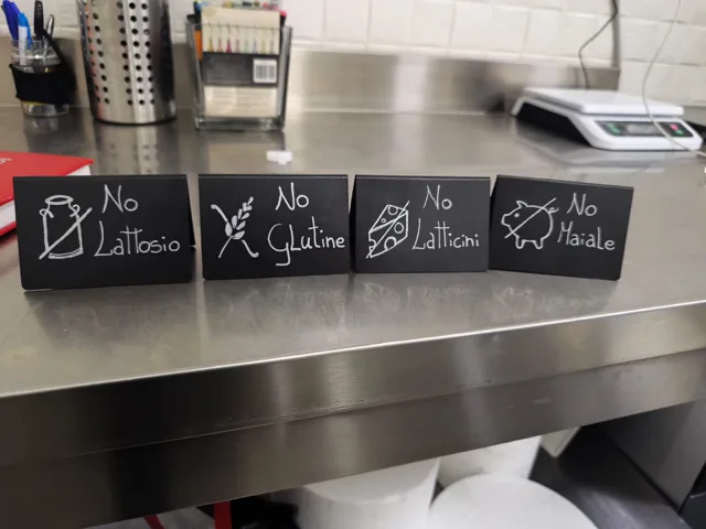 Cartellini neri scritti a mano su un banco inox per indicare preferenze o esclusioni alimentari, con diciture come assenza di lattosio, glutine, latticini e carne di maiale. L'immagine comunica attenzione organizzativa e cura del servizio verso esigenze alimentari diverse.