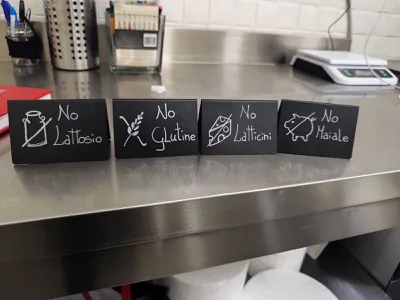 Cartellini neri scritti a mano su un banco inox per indicare preferenze o esclusioni alimentari, con diciture come assenza di lattosio, glutine, latticini e carne di maiale. L'immagine comunica attenzione organizzativa e cura del servizio verso esigenze alimentari diverse.