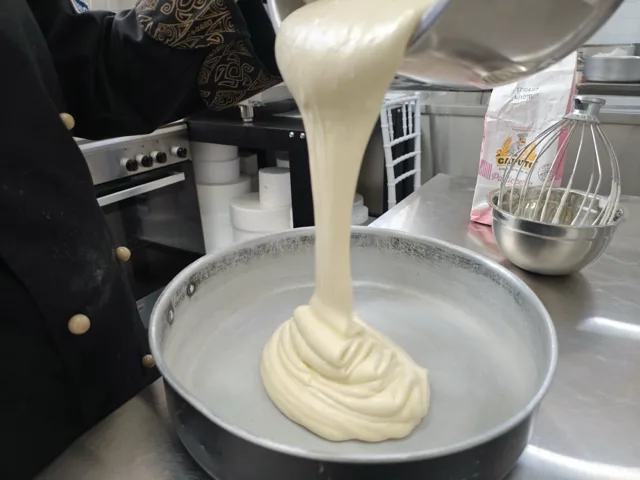 Impasto chiaro e denso versato in una tortiera durante una preparazione di pasticceria, con frusta, ciotola e farina sul banco di lavoro. La foto mostra una fase artigianale del laboratorio dolci di Viglianisi Catering.