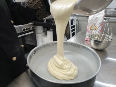Impasto chiaro e denso versato in una tortiera durante una preparazione di pasticceria, con frusta, ciotola e farina sul banco di lavoro. La foto mostra una fase artigianale del laboratorio dolci di Viglianisi Catering.
