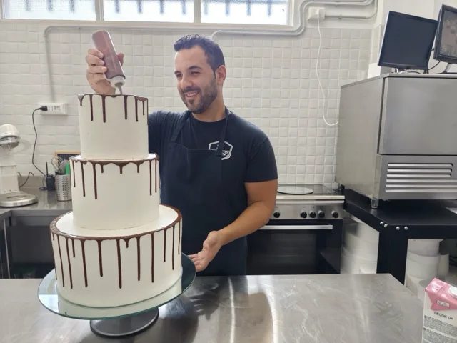 Pasticcere al lavoro su una drip cake bianca a tre piani, mentre applica la colatura di cioccolato lungo il bordo superiore. L'immagine valorizza la cura artigianale della pasticceria firmata Viglianisi Catering.